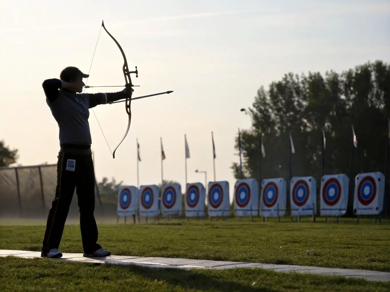 A photograph of archers of varying ages and skill levels participating in a training session at the TAC-TIR archery range, focusing on proper form and technique under the guidance of an instructor.